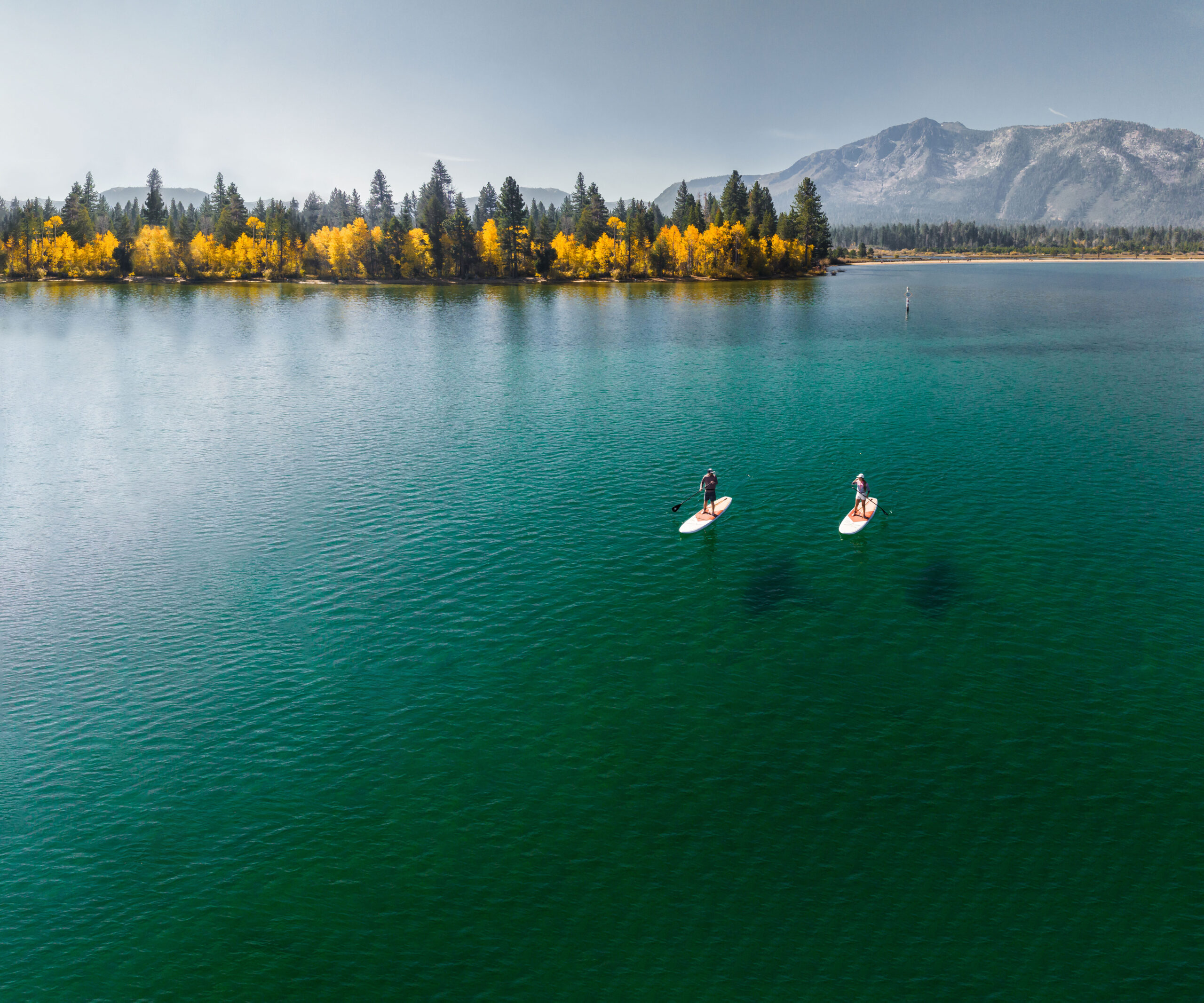 Two Paddleboarders on a blue lake with trees and mountains in the back